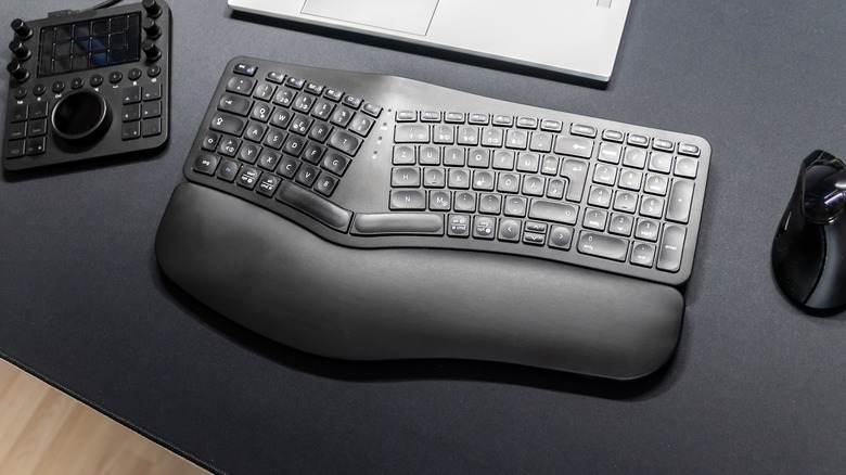 A black, split ergonomic keyboard sits between an ergonomic mouse and a control console on a desk with a black surface, the edge of a silver laptop is just out of frame