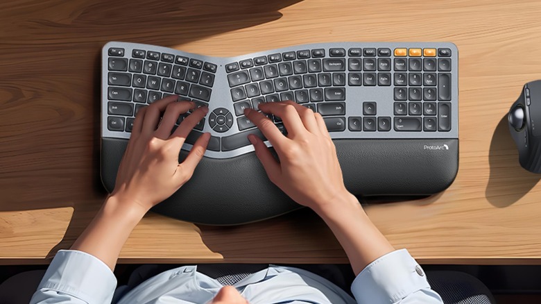 A user sits in front of a ProtoArc EK01 ergonomic keyboard, a black mouse is next to the keyboard, and the user has both hands on the keys
