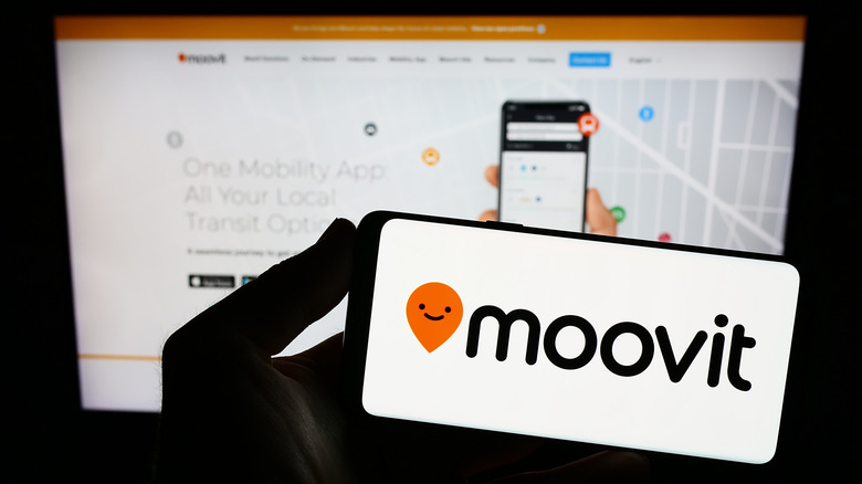 A hand holding an iPhone in a dark room, the Moovit logo is prominent on the iPhone screen.