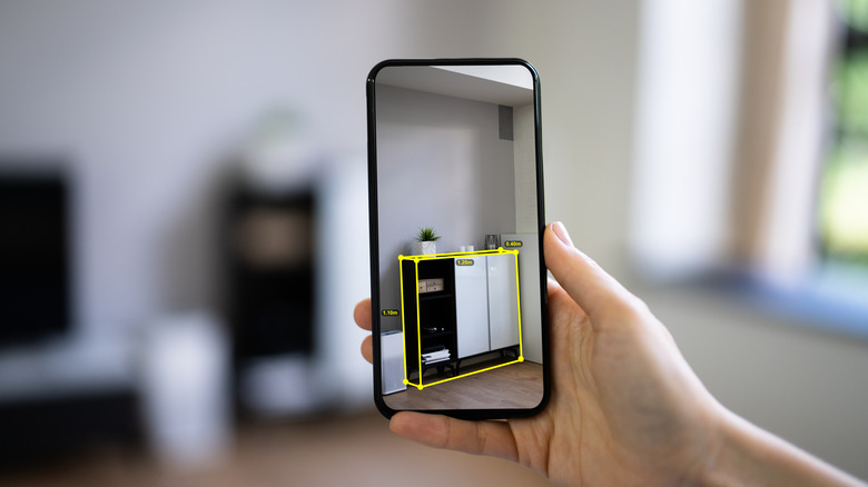 a hand holding a phone with the camera app open to measure furniture in a room