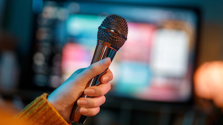 A hand holding a microphone with a TV in the background