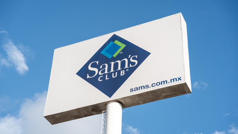 A white sign displaying the Sam's Club logo, the sign has a blue sky and clouds behind it