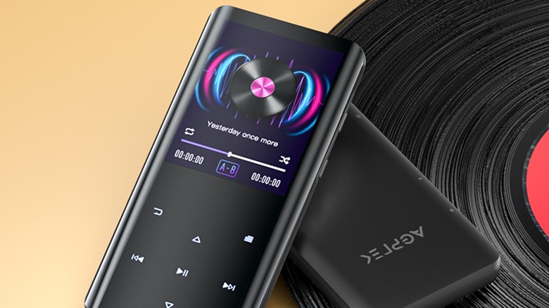 Two black AGPTEK 32GB MP3 Player sitting on top of a vinyl record against a yellow backdrop, one device is face down, whereas the other is face up and showing song information on its screen