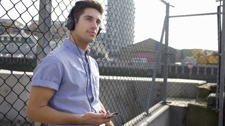 An individual in a blue shirt stand outside against a fence while holding an MP3 player and wearing black headphones, the individual has a small smile on their face