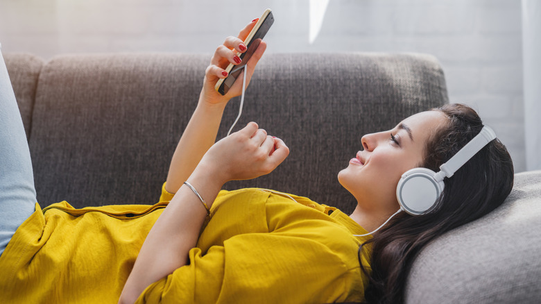 An individual in a yellow shirt lays on a gray couch as they navigate an MP3 player in their right hand, they are also wearing white headphones