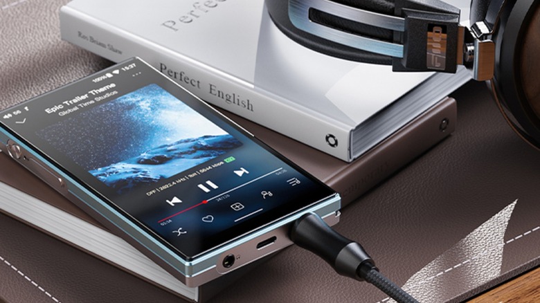 A silver/blue JadeAudio/FiiO JM21 Hi-Res Music Player sits on top of a book as it has a pair of black headphones connected to its aux port, a book titled "Perfect English" sits next to the device