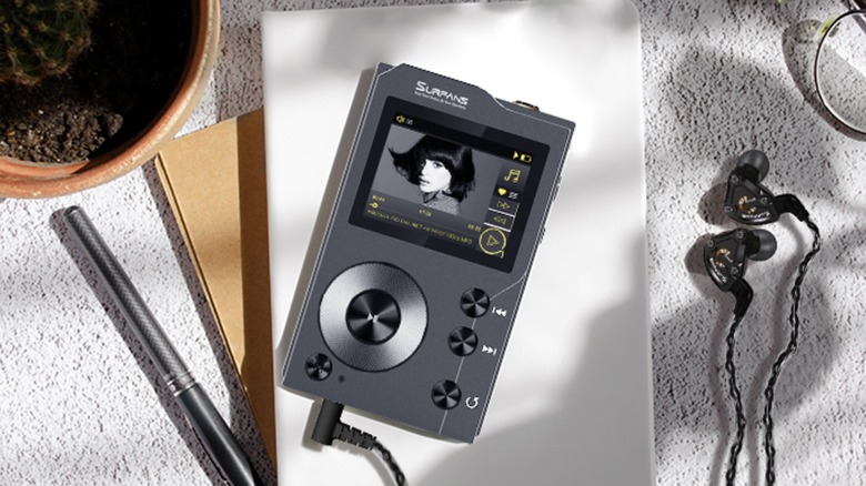 A black Surfans F20 Hi-Fi MP3 Player displays song information on its display as it sits on top of a white book, next to it is a pair of headphones, a potted plant, a pen, and a pair of glasses