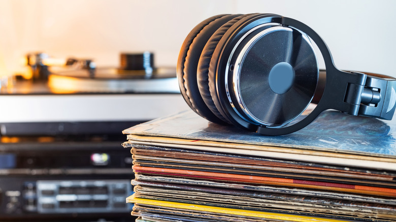 A pair of black headphones sits on top a pile of vinyl records, an audio setup with a turntable on top if blurred slightly in the background