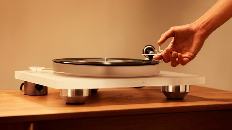A clear Marantz TT-15S1 Manual Belt-Drive Premium Turntable sits on a table, an individual's right hand is coming into frame to move the tonearm of the unit