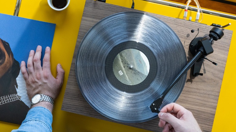 A Pro-Ject Debut Evo 2 Turntable with a woodgrain finish sits on a bright yellow countertop, an individual's right hand is adjusting the tonearm to find a specific location on the vinyl record