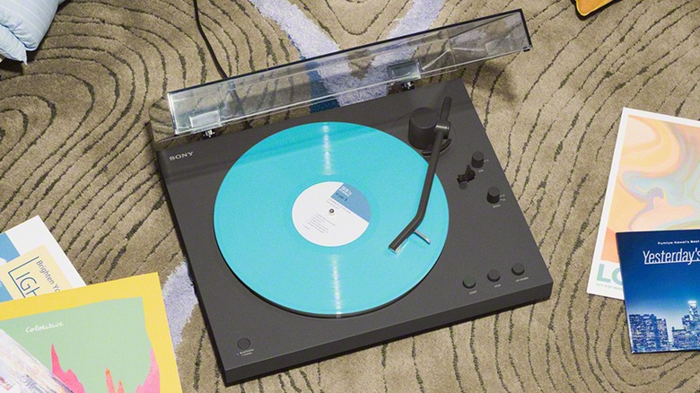 A black Sony PS-LX3BT Wireless Bluetooth Turntable sits on patterned carpet next to several records, a neon blue vinyl sits on the record player and the tonearm is about halfway through the record
