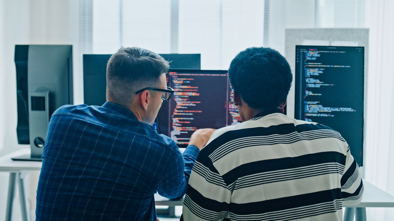 Two people collaborating at a desk with code displayed on multiple monitors.