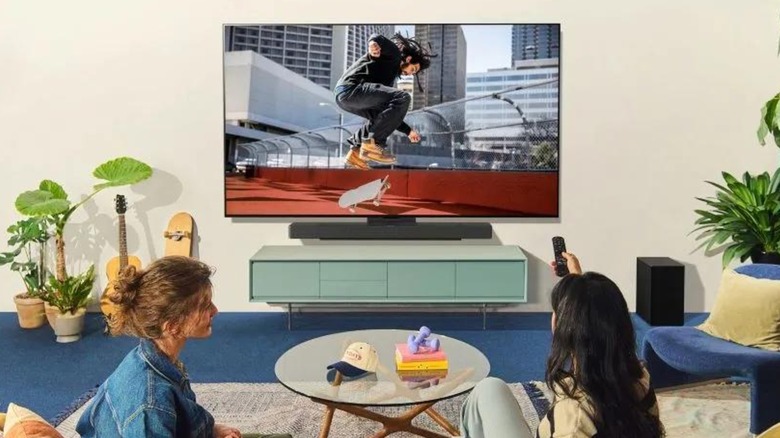 Two individuals are seated in front of the LG 77-inch Class C5 Series OLED Smart TV, with one of the individuals using their right hand to point the controller at the TV