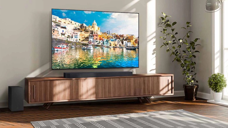 A TV sits above the Samsung QS730D 3.1.2 Channel Soundbar, which sits on a brown TV stand, two plants also appear in the photo