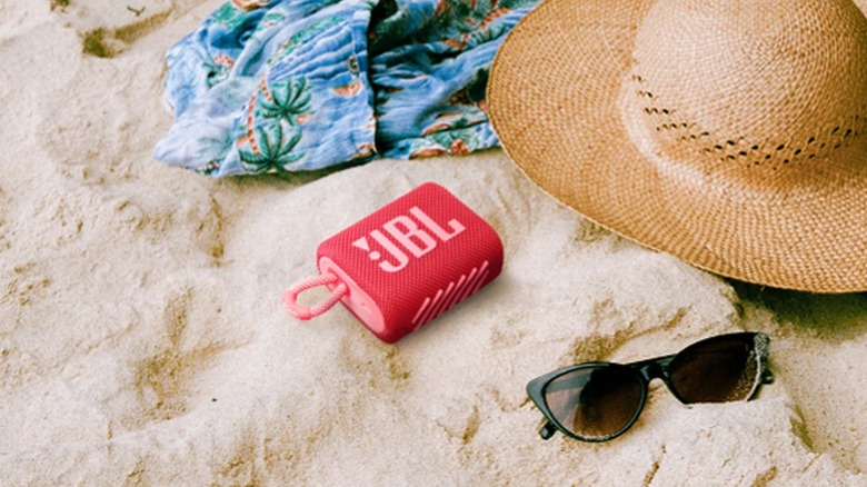 A red JBL Go 3 Portable Mini Bluetooth Speaker lays in sand next to a pair of sunglasses, a sun hat, and a tropical-themed garmet