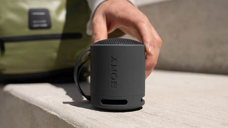 The right hand of an individual is placed on top of a black Sony SRS-XB100 portable speaker