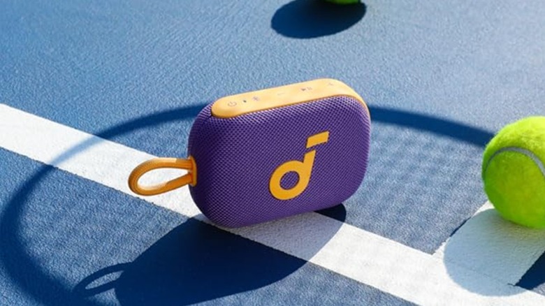 A purple Soundcore Select 4 with yellow trim sits on a tennis court next to a tennis ball