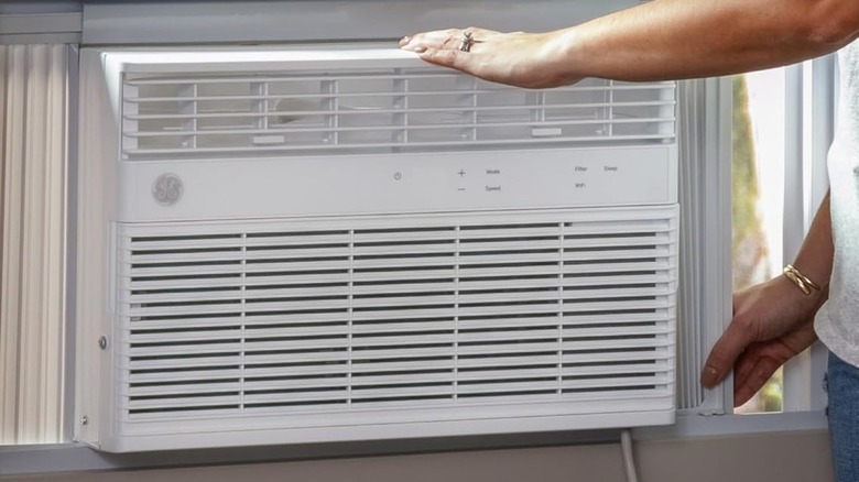 A white GE Window Air Conditioner is being installed in a white windowsill, the arms of an individual can be seen as they have one hand on the top of the unit and another on the side of the unit