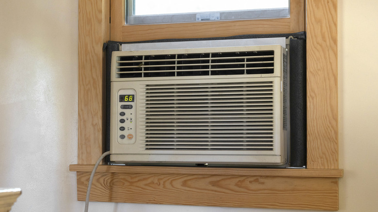 A white air conditioner sits in a windowsill that has brown framing around the window