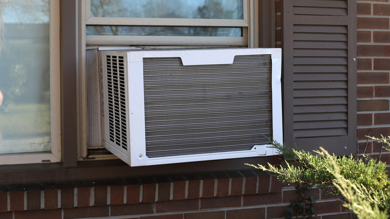 A white window Air Conditioning unit is installed in a window of a brown brick house