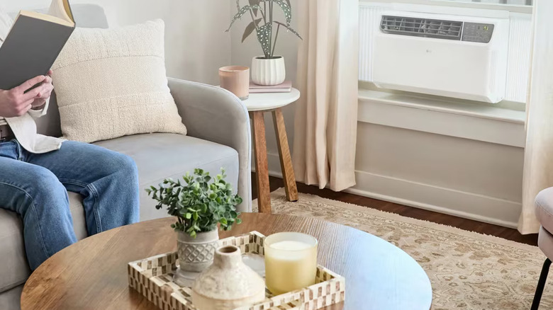A white LG Dual Inverter Smart Air Conditioner inside a home, a couch, coffee table, corner table, and in individual in a white shirt and blue jeans reading a book can all be seen