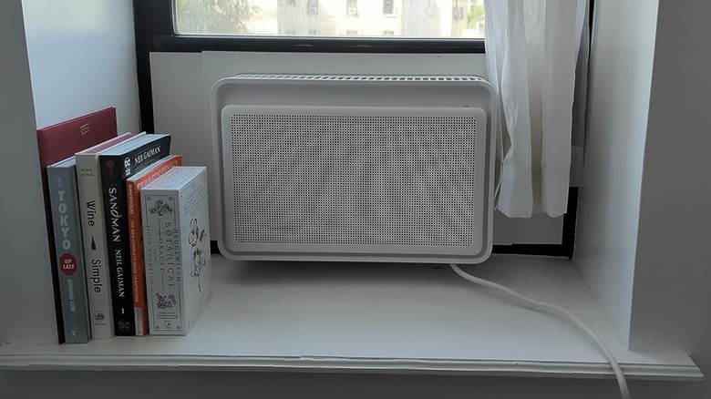 A white Windmill 6,000 Window Air Conditioner sits in a windowsill that has its own cubby, there are several books sitting next to the unit