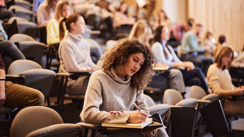 A girl sitting at a desk in a lecture hall with other students in the background behind her