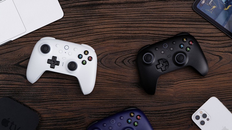 Blue, white, and black 8BitDo controllers on a wooden table surrounded by gaming platforms