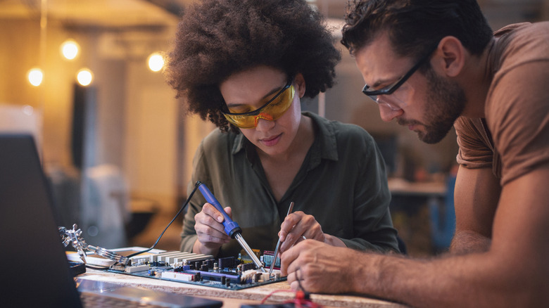 Two people soldering components on a circuit that's placed on a table