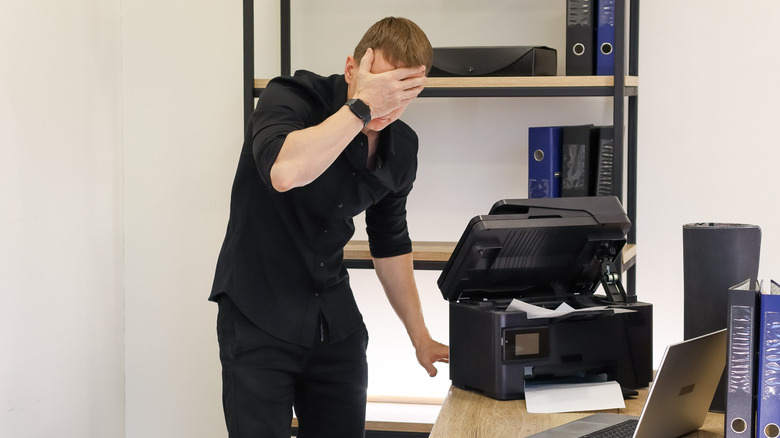 A person covers their face with a hand in frustration after dealing with printer issues in an office environment