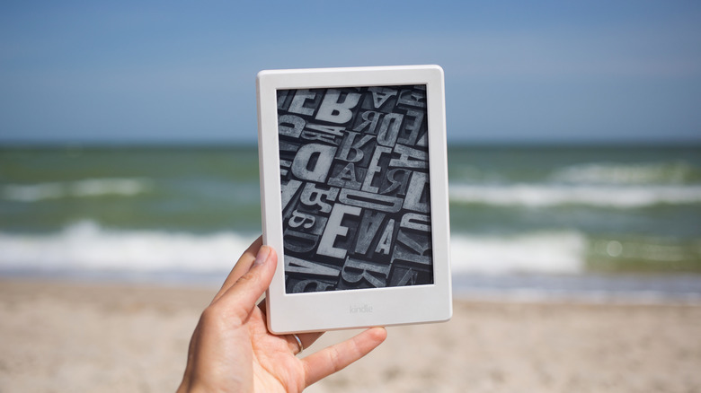 Kindle e-reader held up on a beach with the ocean in the background.