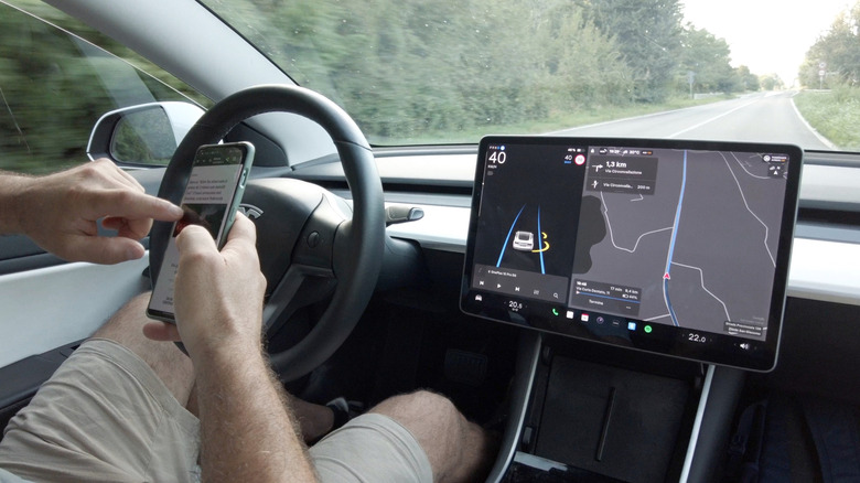 Tesla driver using a smartphone while the center screen shows self-driving navigation and lane visuals.