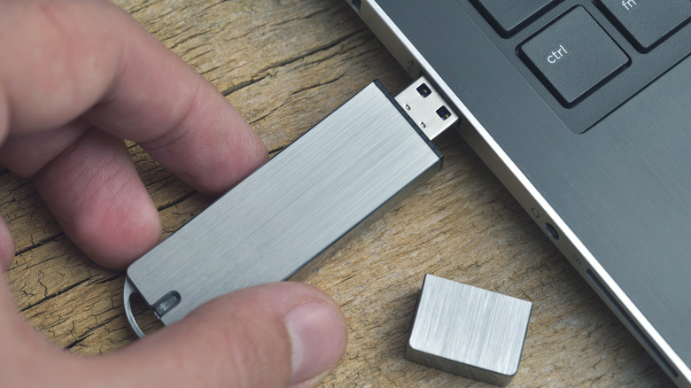 A hand plugging in a metal USB drive into a laptop on a wooden table