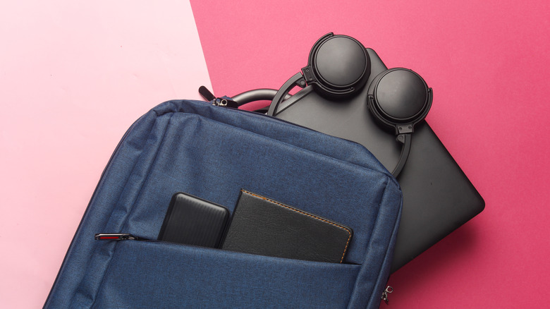 A dark blue backpack with a leather note pad and electronic device in the front pocket, a black laptop and a pair of headphones are underneath