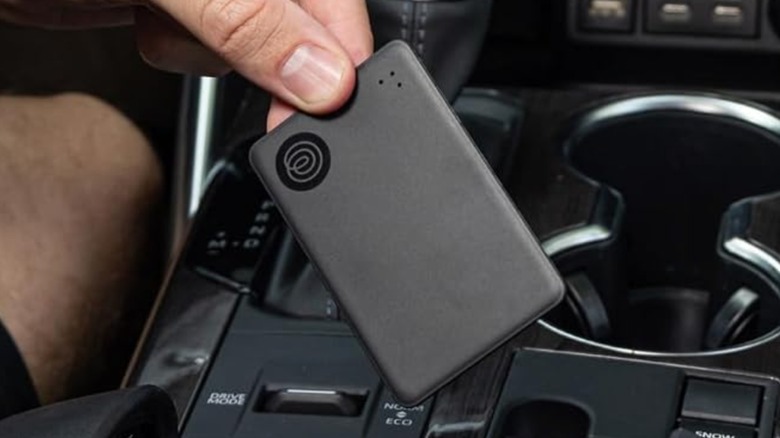 A black Life360 Tile Slim Bluetooth Tracker being held in a user's left hand next to the shifter in a vehicle