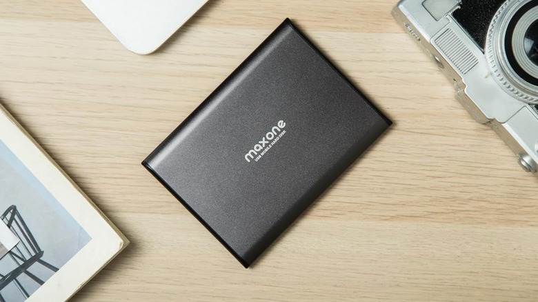 A black Maxone Ultra Slim Portable External Hard Drive sits on a table next to a camera, and what appears to be a book and a laptop