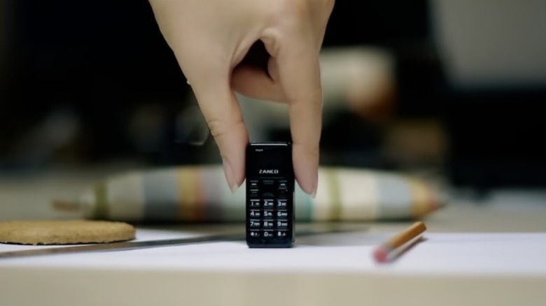 A black Zanco Tiny T1 standing upright on a table next to a pen and a coaster as an individual holds the phone between their thumb and index finger