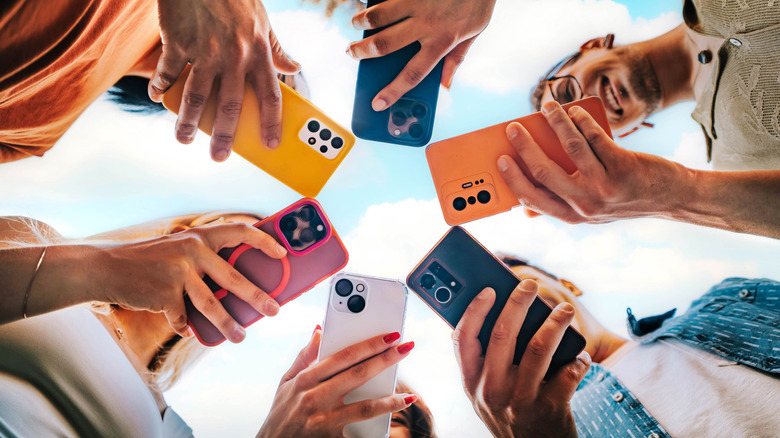 Multiple people standing in a circle, the photo is taken from the ground level to look up at the individuals as they hold smartphones in a circular pattern, the smartphones are in protective cases and it makes for a colorful display