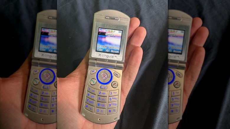 A photo of an individual's left hand as it holds a silver Pantech C300 flip-phone, the phone is displaying its Home screen on its tiny display
