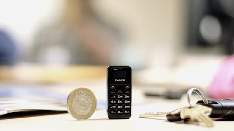 A black Zanco Tiny T1 cellphone standing upright on a white table next to a coin and a set of car keys, the coin is also standing upright
