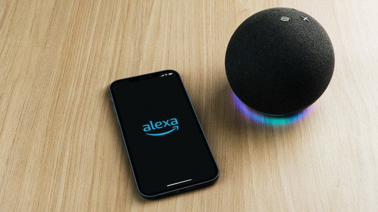 An black Amazon Echo device sits next to a smartphone displaying the Alexa logo on its screen, both items are sitting on a wooden counter