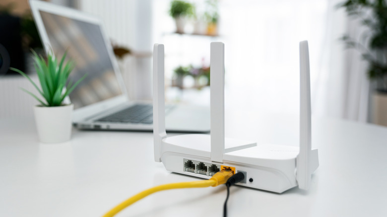 A white router sitting on a white table, a MacBook and potted plant are seen blurred in the background