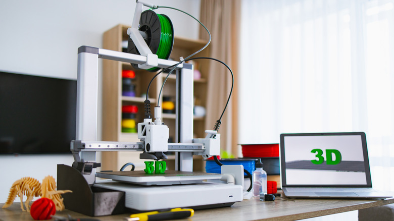 3D printer placed alongside a laptop on a wooden table next to a glass door