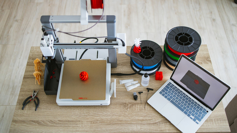 3D printer placed on a large wooden table alongside a laptop and spools of filament