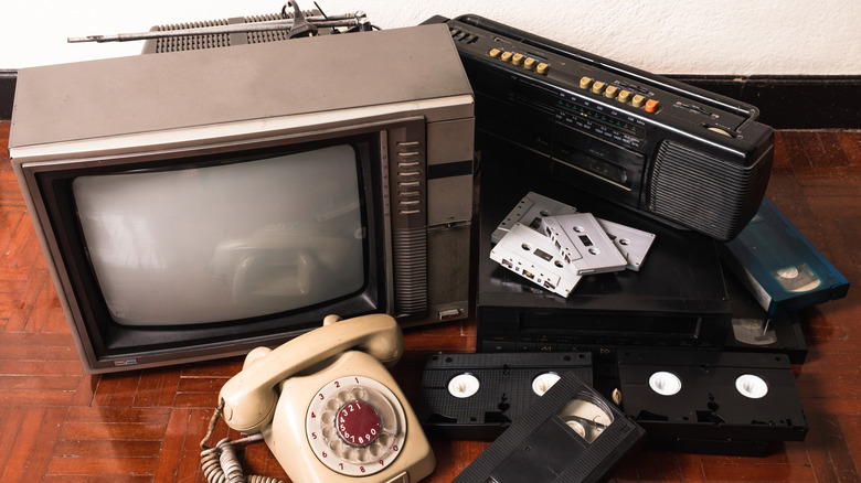 A pile of old technology, including a tube TV, a rotary phone, VHS tapes, and a boombox.