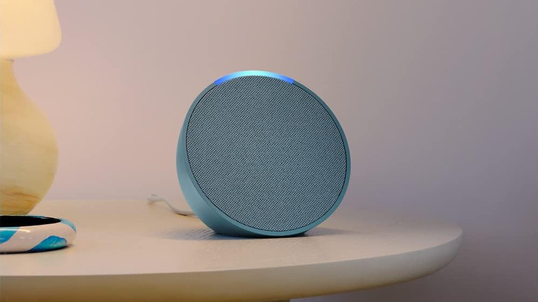 Amazon Echo Pop smart speaker sitting on a bedside table beside a lamp.
