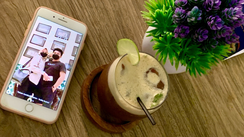 An iPhone 8 on a table next to a soft drink and a vase of flowers