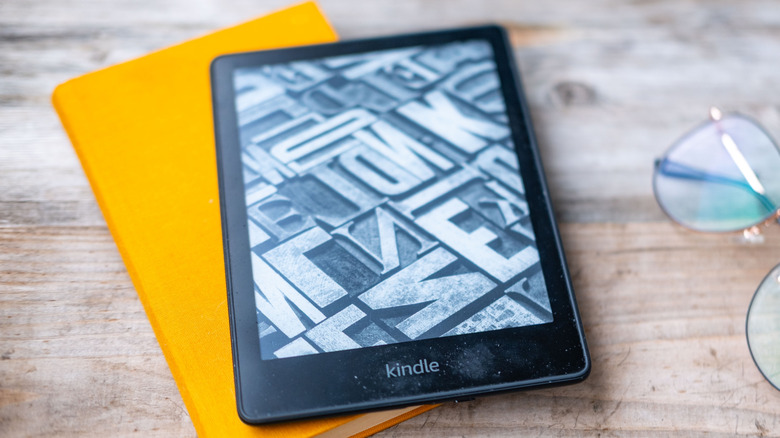 Kindle e-reader resting on a yellow book beside a pair of eyeglasses on a wooden table.