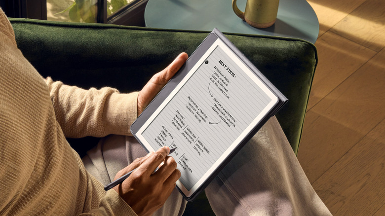 Person writing notes on a Kindle Scribe with a stylus while sitting on a couch.