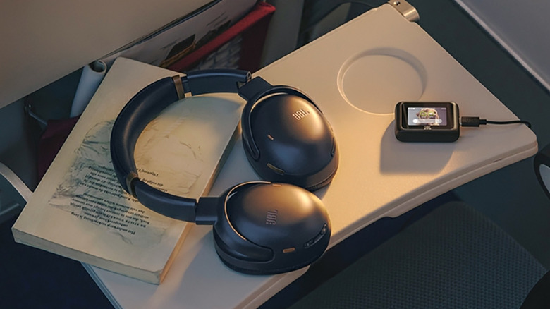 The JBL Tour One M3 Smart Tx headphones atop an airplane tray with a media player and book.